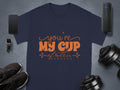 A navy blue graphic T-shirt by Garment Graphics features Youre My Cup of Tea in bright orange script. Made from 100% cotton, it’s displayed with a smartphone, smartwatch, wireless speaker, shoes, dumbbells, and headphones on a sleek gray background.