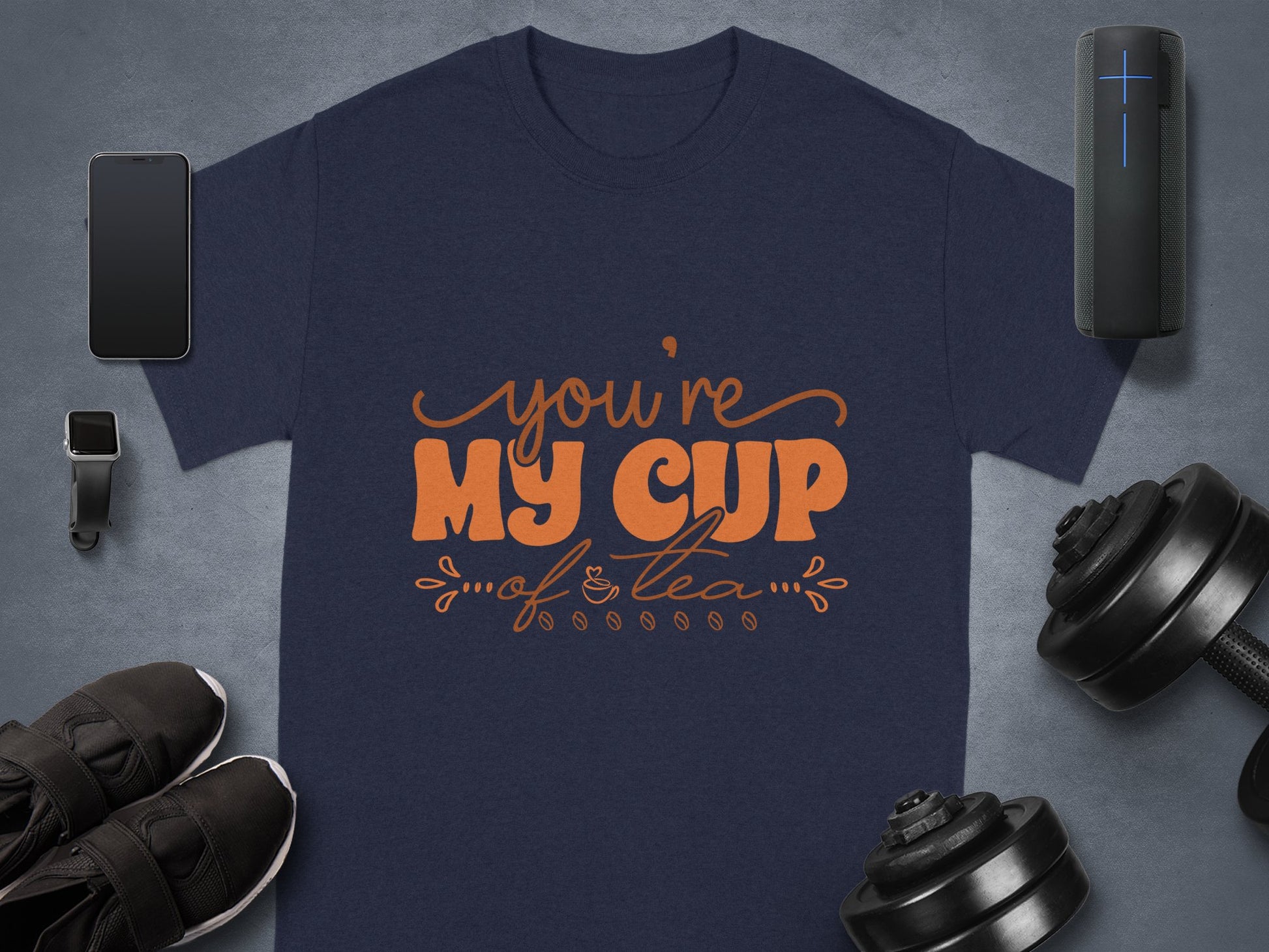 A navy blue graphic T-shirt by Garment Graphics features Youre My Cup of Tea in bright orange script. Made from 100% cotton, it’s displayed with a smartphone, smartwatch, wireless speaker, shoes, dumbbells, and headphones on a sleek gray background.