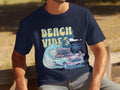 A person relaxes on a wooden bench wearing Garment Graphics navy blue graphic t-shirt adorned with BEACH VIBES in yellow, showcasing a classic car, surfboard, large wave, and palm trees—capturing a retro beach theme.