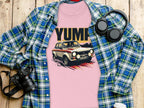 A pink Garment Graphics tee featuring a vintage car graphic and YUMP lies on wood, layered with a blue plaid shirt. Nearby are a camera, headphones, and a white coffee cup.