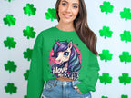 A woman beams, dressed in a vibrant Garment Graphics sweatshirt featuring a unicorn with I love my pony. Her long brown hair cascades over denim shorts as she stands against green shamrocks, exuding playful charm and whimsical style.