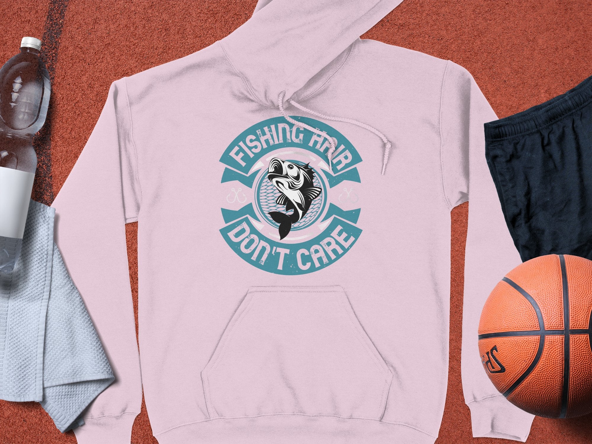 A light pink Garment Graphics hoodie with Fishing Hair Dont Care surrounds a fish design on a sports surface, perfect for anglers. Nearby essentials include a basketball, black shorts, water bottle, and towel for relaxation or play.