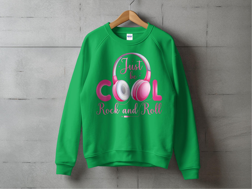 A green sweatshirt from Garment Graphics, displayed on a wooden hanger against a gray wall, showcases the Rock and Roll Pink Headphones graphic. The Just be Cool, Rock and Roll! slogan in pink and white makes it a trendy pick for music fans.