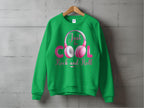 A green sweatshirt from Garment Graphics, displayed on a wooden hanger against a gray wall, showcases the Rock and Roll Pink Headphones graphic. The Just be Cool, Rock and Roll! slogan in pink and white makes it a trendy pick for music fans.