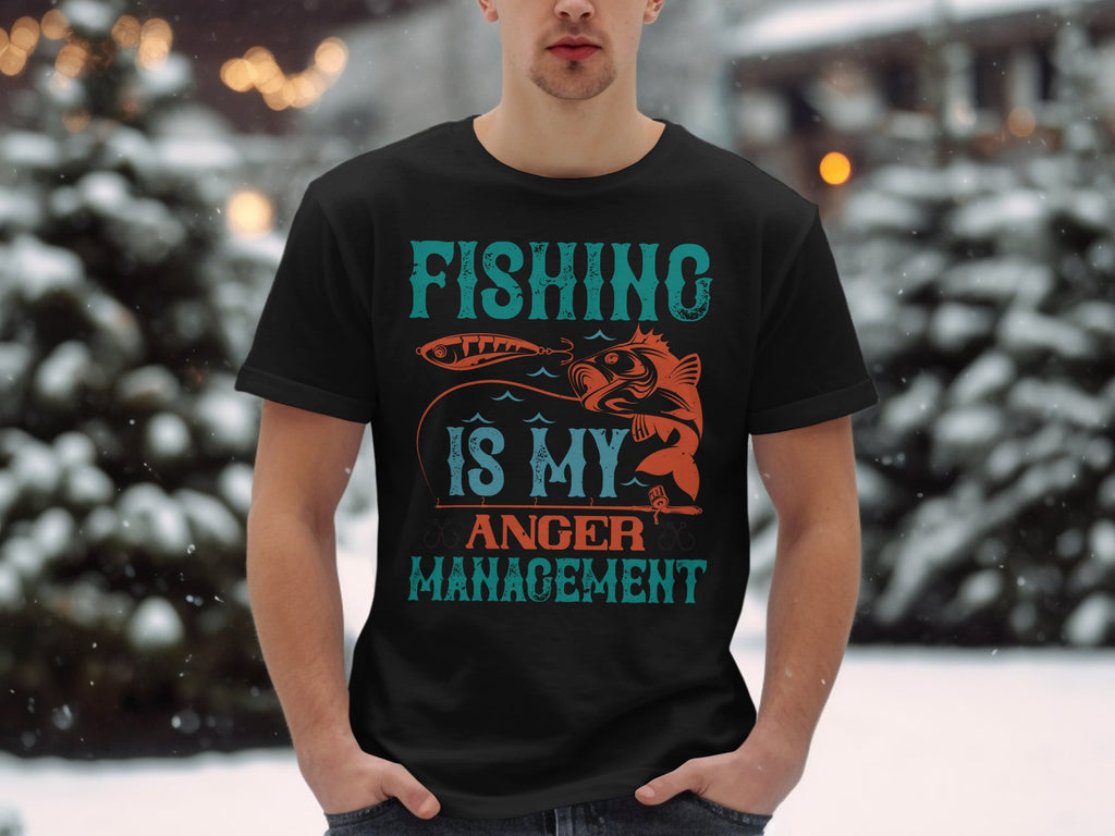 A fishing enthusiast sports a Garment Graphics black T-shirt featuring Fishing is my anger management in bold blue and orange text, standing against a serene snowy landscape with blurred evergreen trees.