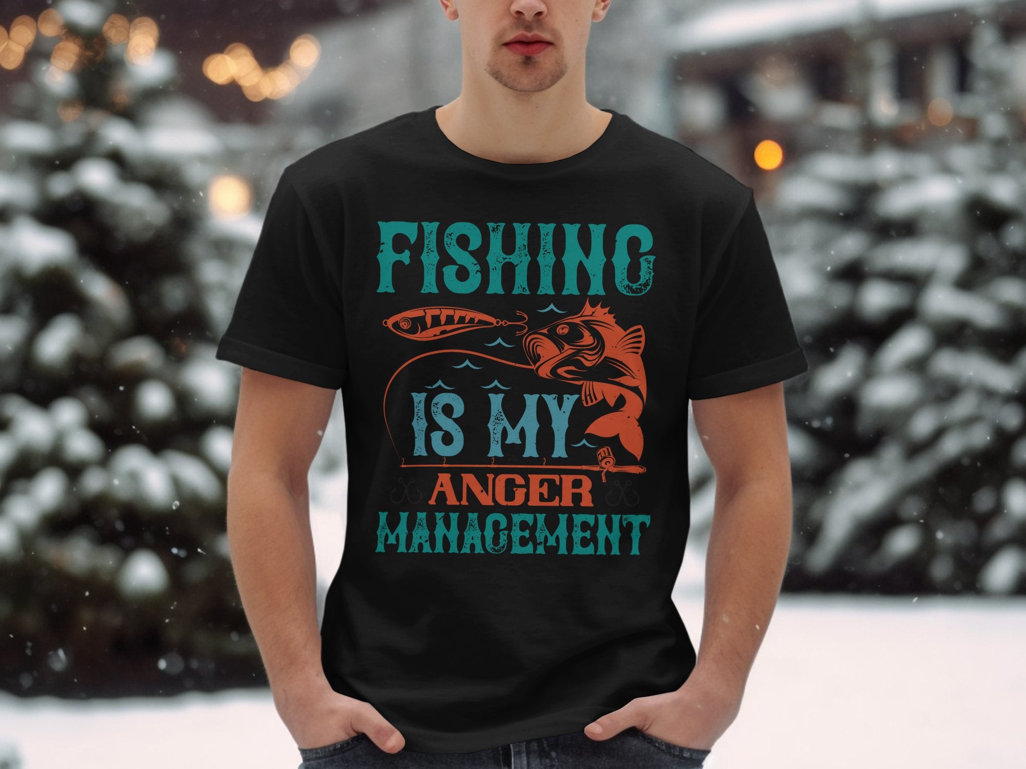 A fishing enthusiast sports a Garment Graphics black T-shirt featuring Fishing is my anger management in bold blue and orange text, standing against a serene snowy landscape with blurred evergreen trees.