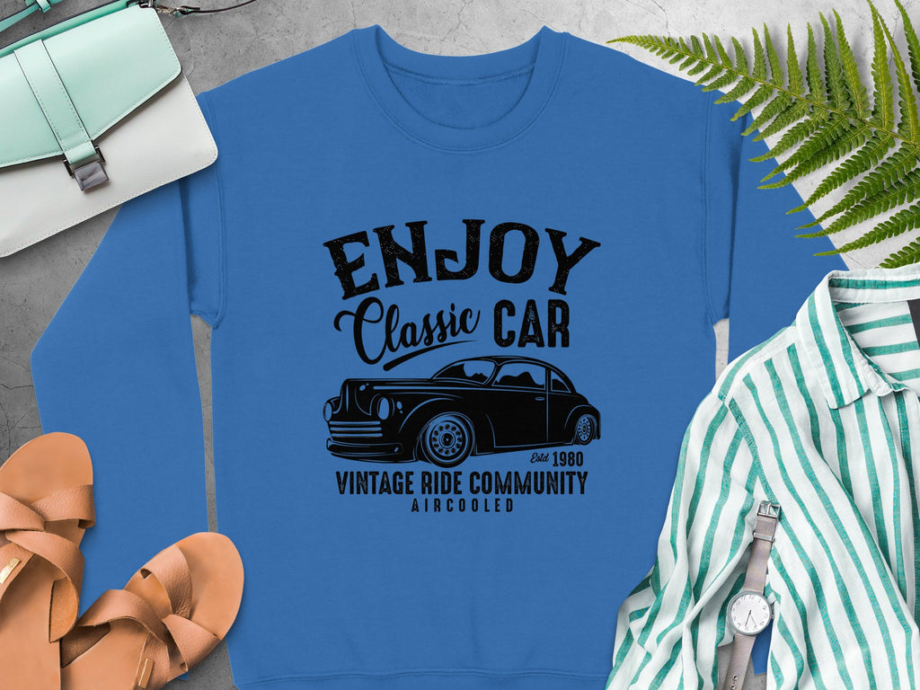 Garment Graphics offers a blue sweatshirt with a black graphic of a classic car and the text Enjoy Vintage Ride, Aircooled, Estd 1980. Its stylishly combined with a purse, sandals, wristwatch, and striped shirt on a gray surface.