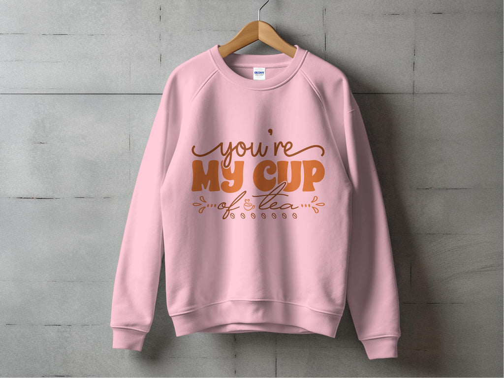 The Garment Graphics sweatshirt, featuring the playful youre my cup of tea in vibrant orange, hangs stylishly on a wooden hanger against a concrete wall—ideal for tea lovers who appreciate quirky charm and comfort.