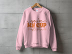 The Garment Graphics sweatshirt, featuring the playful youre my cup of tea in vibrant orange, hangs stylishly on a wooden hanger against a concrete wall—ideal for tea lovers who appreciate quirky charm and comfort.
