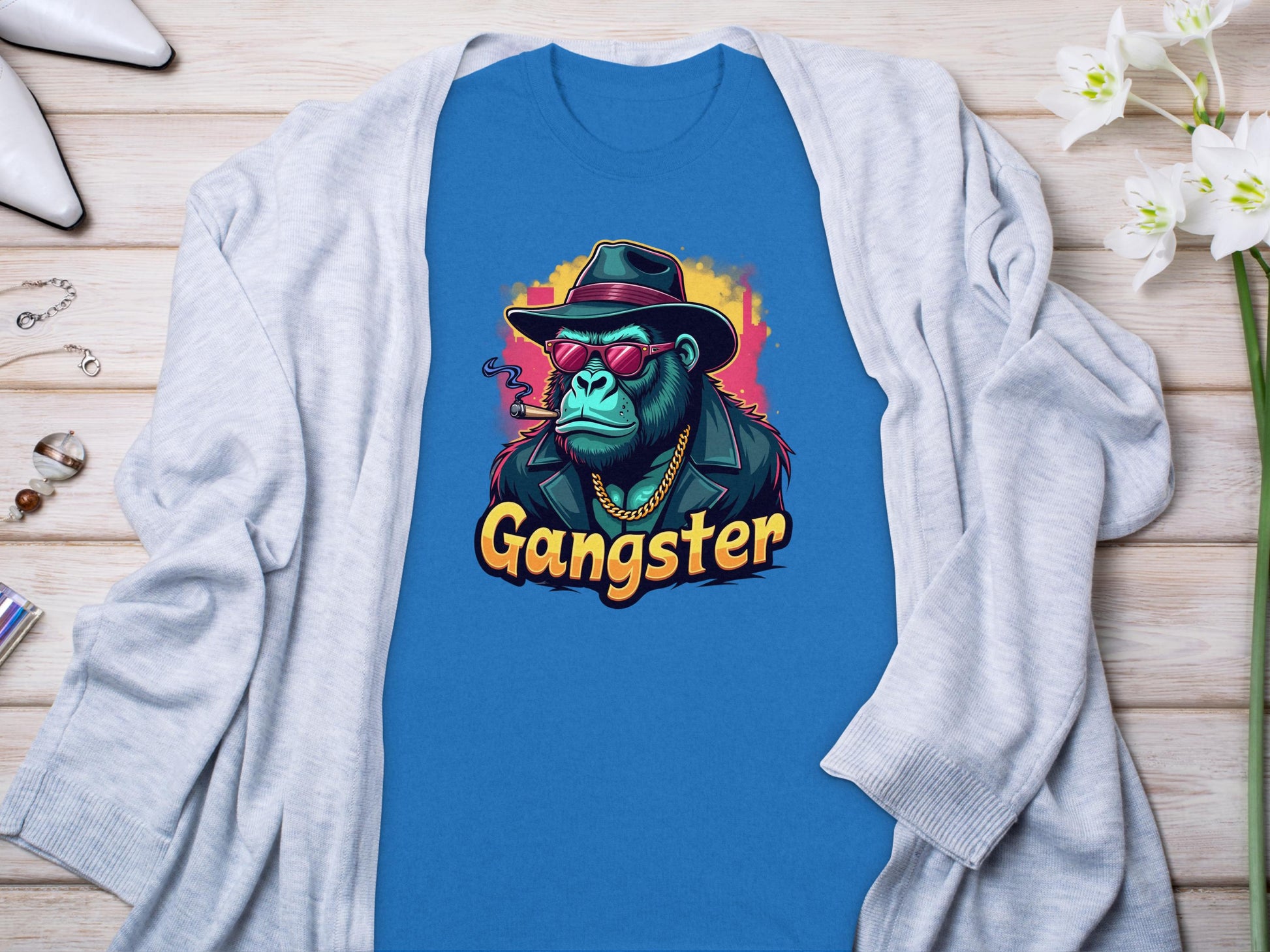 The Garment Graphics Gangster Gorilla T-Shirt features a cartoon ape in a hat, sunglasses, gold chain, and black coat with Gangster printed below. Its styled on a light wood surface next to a classic fit cardigan and flowers for an effortlessly cool urban vibe.