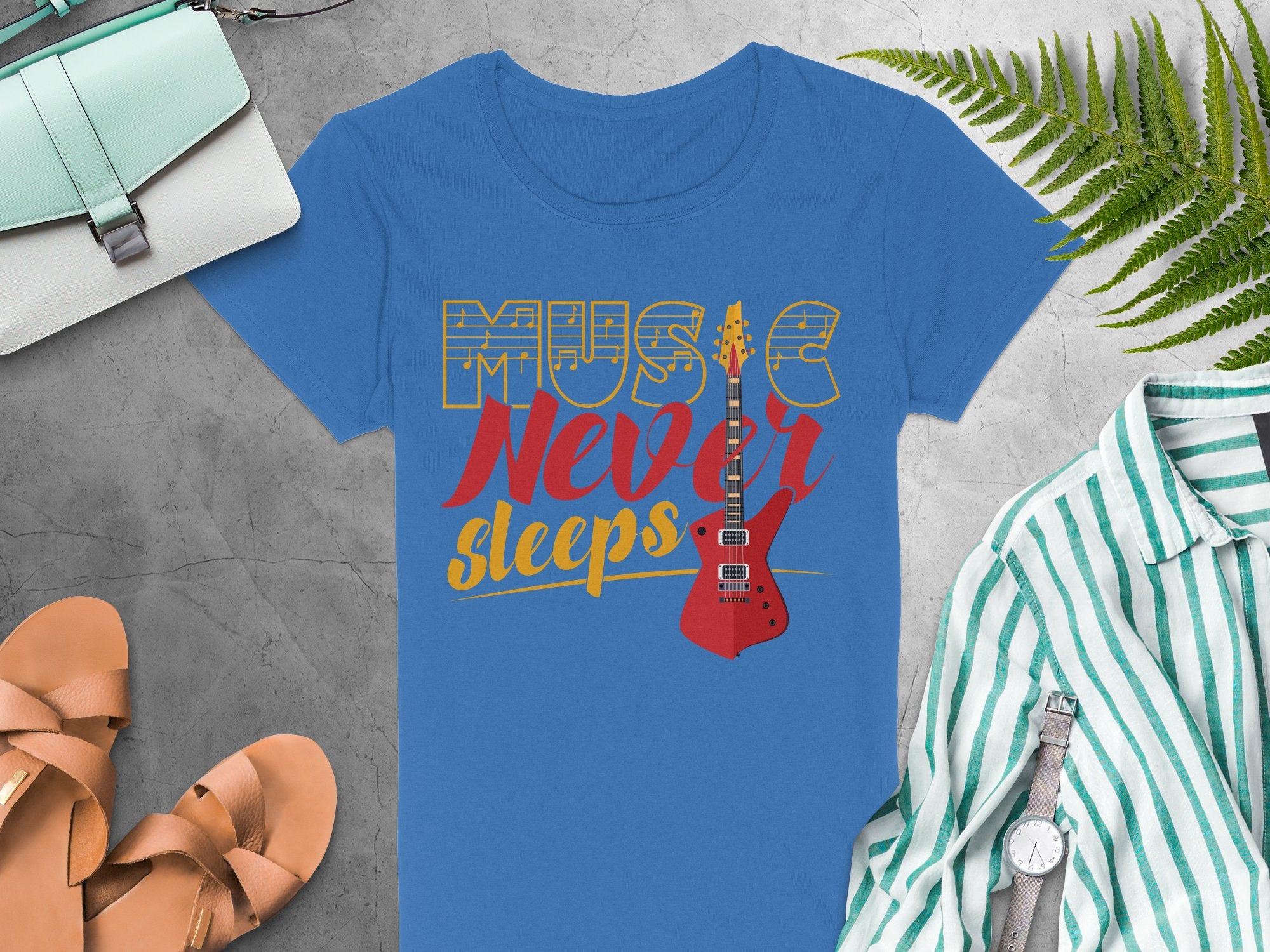 Garment Graphics blue t-shirt, featuring a red electric guitar graphic and bold Music Never Sleeps, offers a striking design for unique tee fans. Styled on gray with a purse, tan sandals, wristwatch, striped shirt, and greenery sprig.