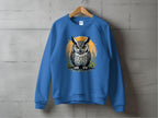 A blue unisex sweatshirt by Garment Graphics, featuring an owl with large eyes amidst yellow foliage against an orange circle, hangs on a wooden hanger against a concrete wall.