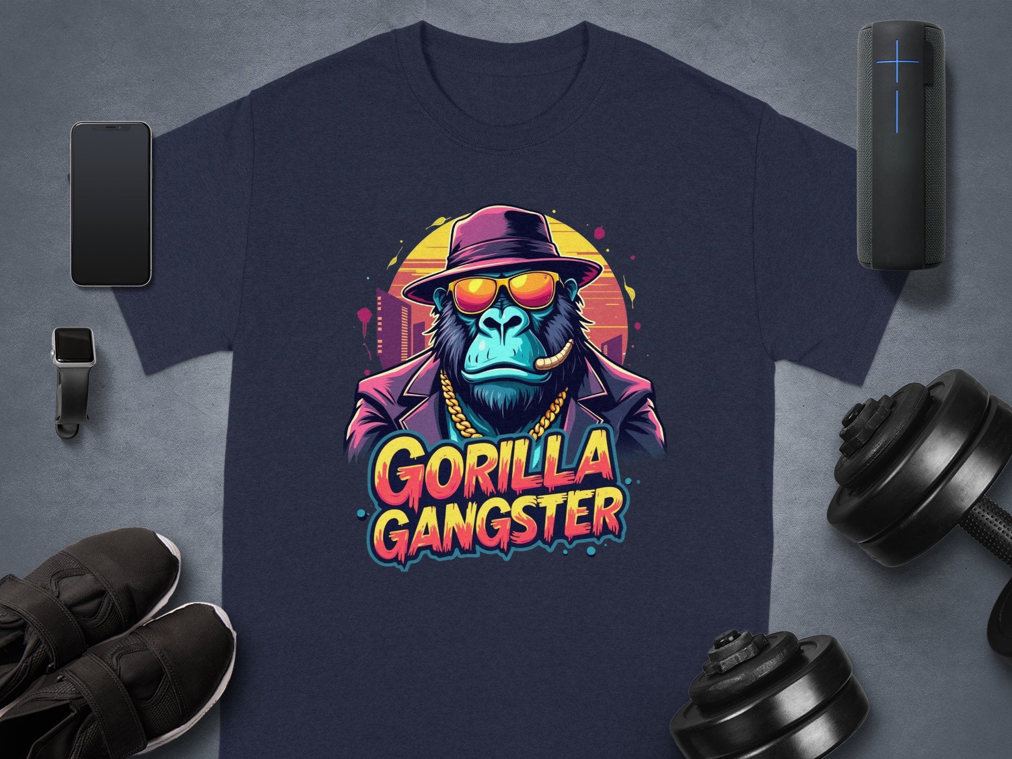 The Garment Graphics navy blue T-shirt features a gorilla in a fedora, sunglasses, and suit enjoying a cigar. GORILLA GANGSTER is printed below in vibrant letters, surrounded by accessories like a phone, smartwatch, shoes, and weights.