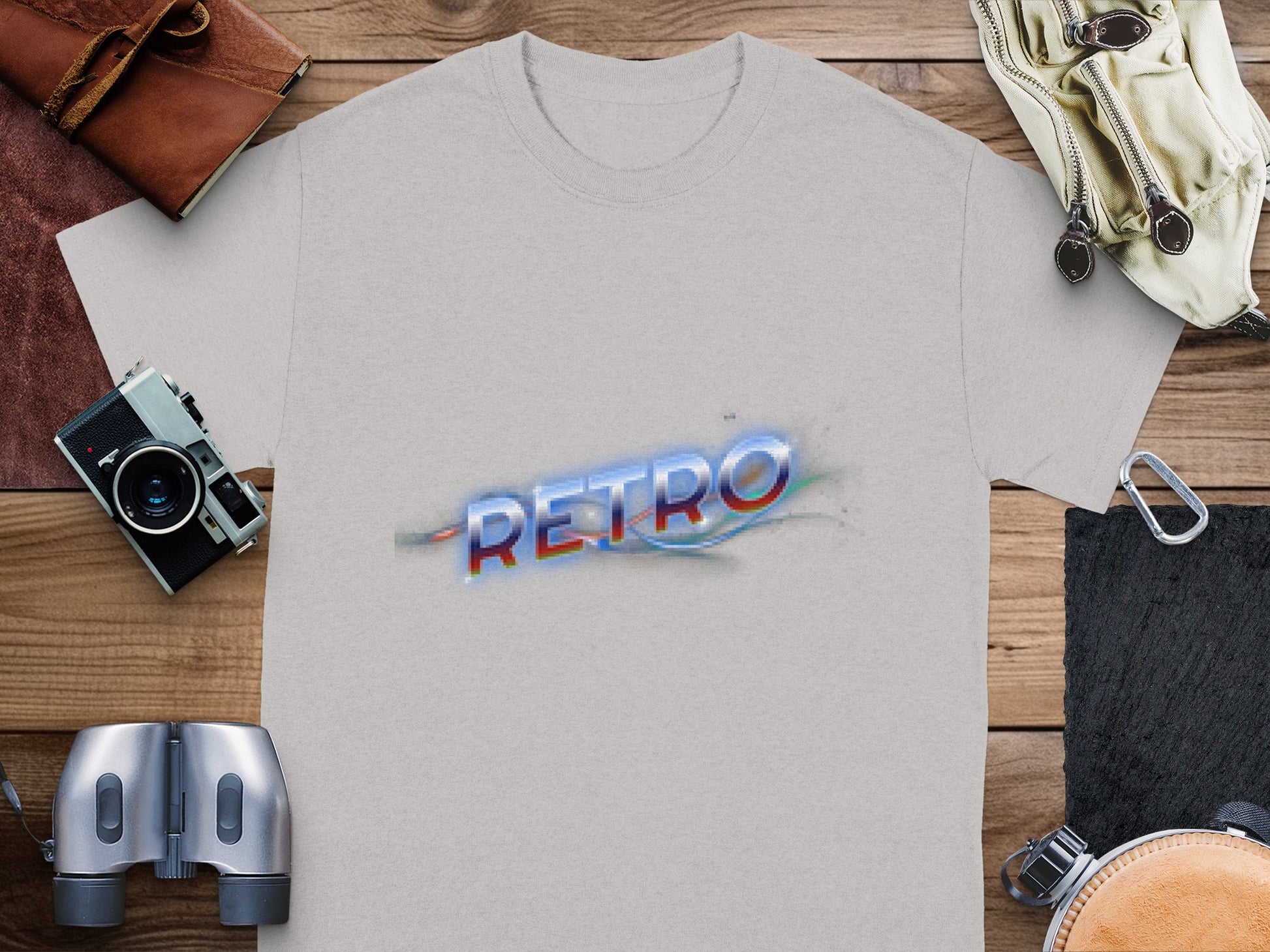 A classic fit gray t-shirt from Garment Graphics, featuring RETRO in colorful, blurred text, lies on a wooden surface surrounded by a leather wallet, canvas bag, vintage camera, binoculars, and metallic buckles for a vintage vibe.
