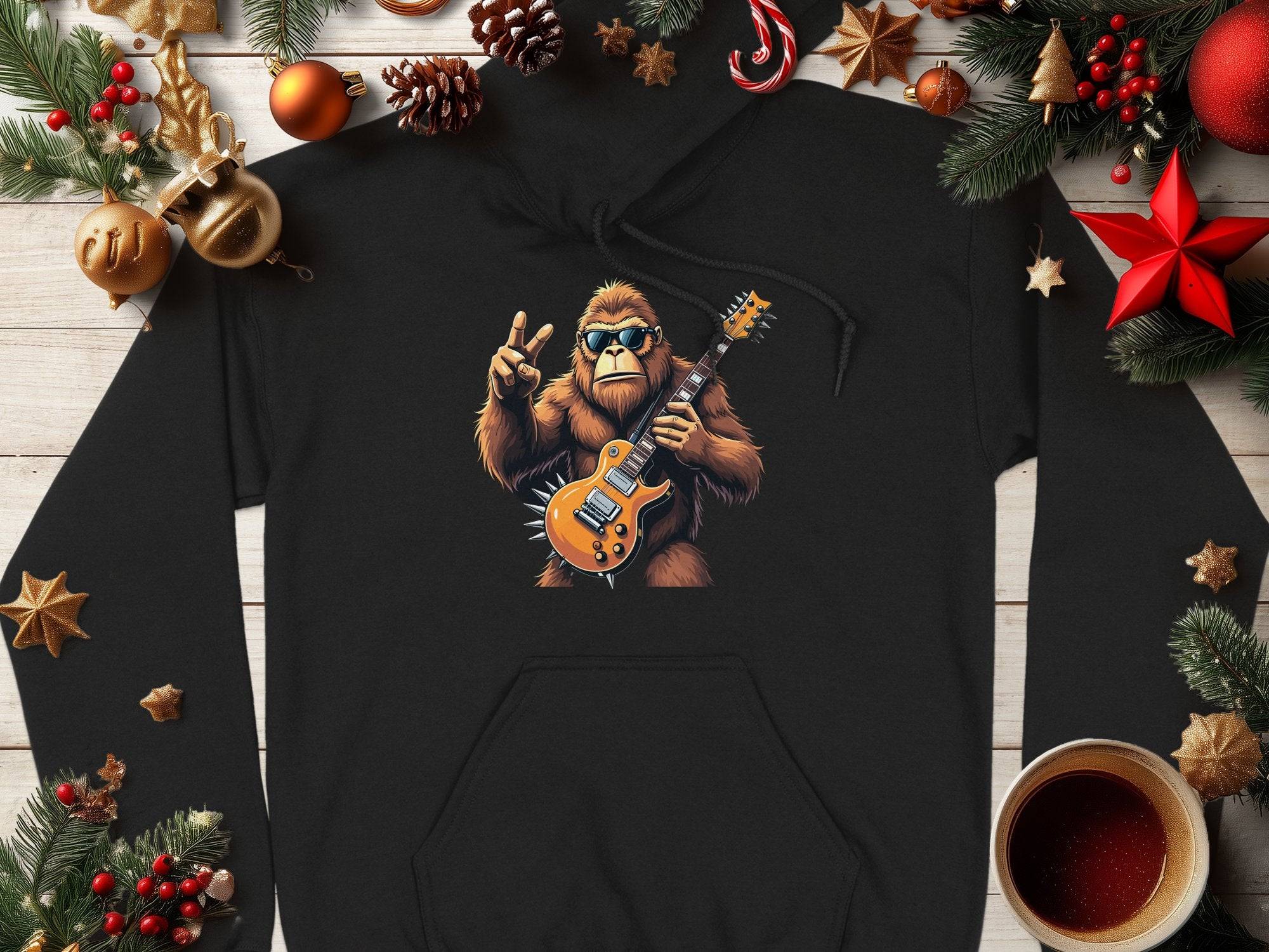 A Garment Graphics black electric guitar hoodie features a yeti playing guitar and a peace sign, surrounded by festive elements like pinecones, red stars, gold ornaments, and a steaming coffee mug on wood. Ideal for music enthusiasts!.