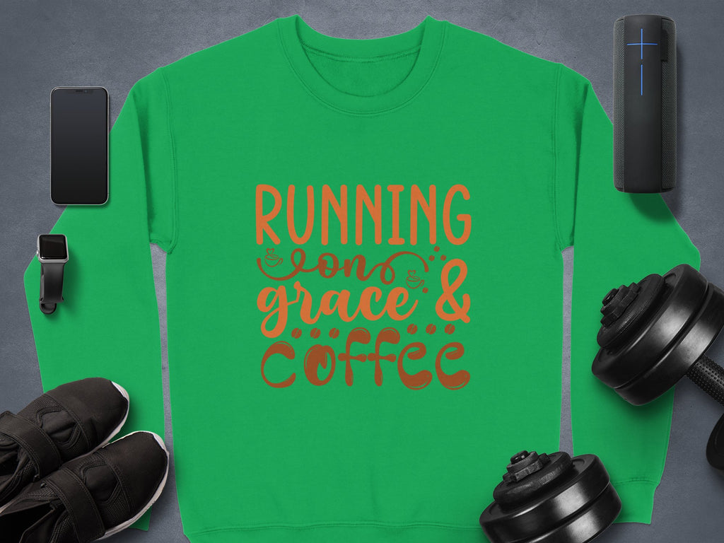 A green Garment Graphics unisex sweatshirt features Running on Grace and Coffee in bold orange letters, surrounded by a smartphone, smartwatch, wireless speaker, sneakers, and two adjustable dumbbells on a dark surface.