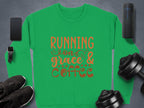 A green Garment Graphics unisex sweatshirt features Running on Grace and Coffee in bold orange letters, surrounded by a smartphone, smartwatch, wireless speaker, sneakers, and two adjustable dumbbells on a dark surface.