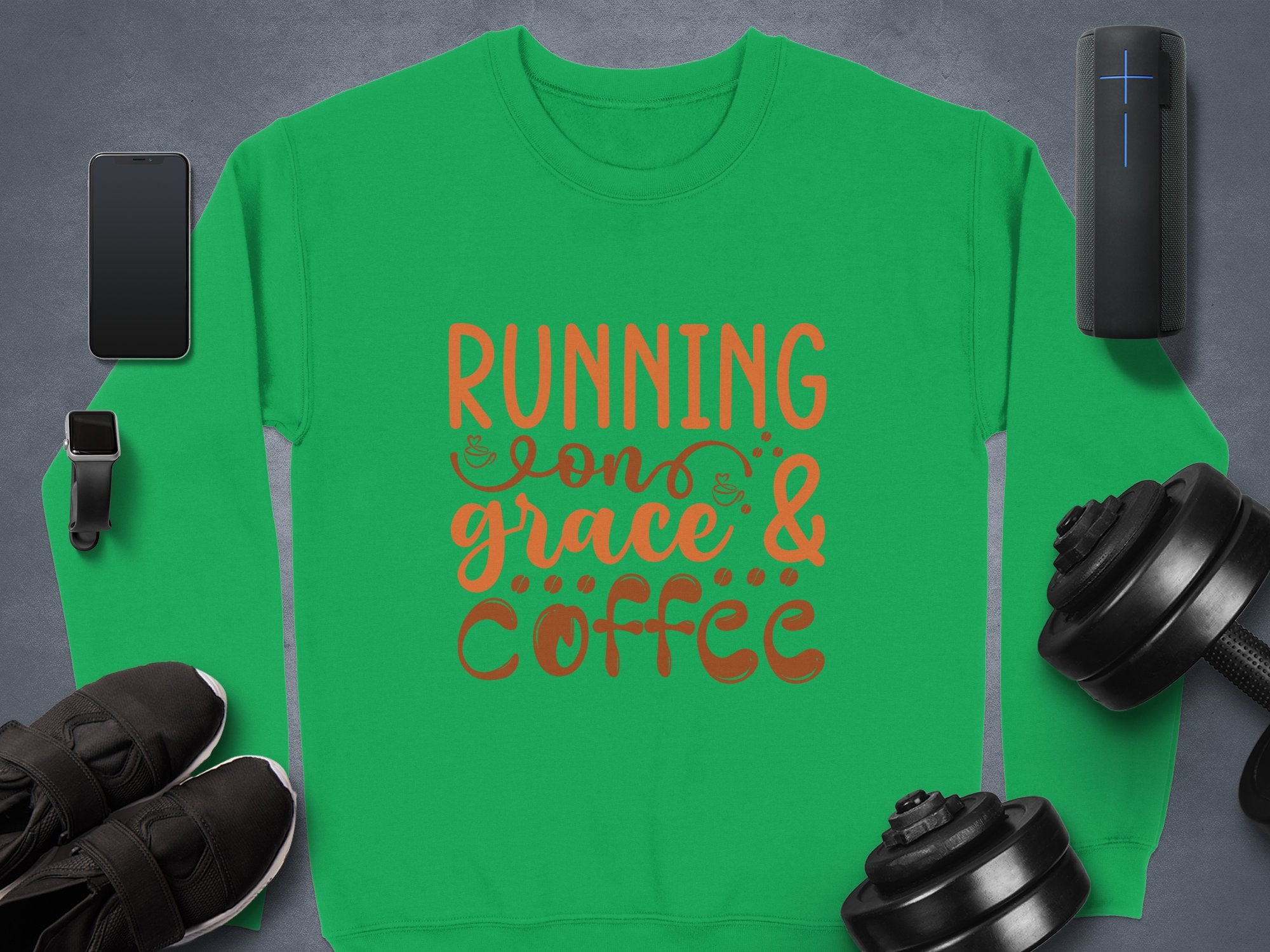 A green Garment Graphics unisex sweatshirt features Running on Grace and Coffee in bold orange letters, surrounded by a smartphone, smartwatch, wireless speaker, sneakers, and two adjustable dumbbells on a dark surface.