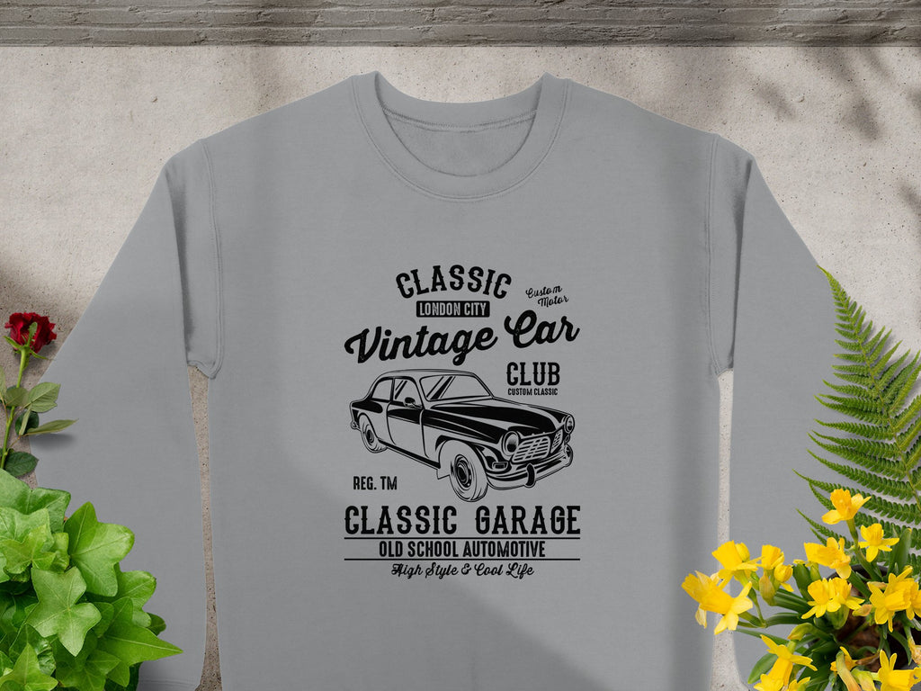 A Garment Graphics gray T-shirt featuring a retro car graphic and text Classic London City Vintage Car Club Custom Garage with High Style & Cool Life is displayed on a flat surface, accented by yellow flowers and green leaves to highlight its automotive flair.