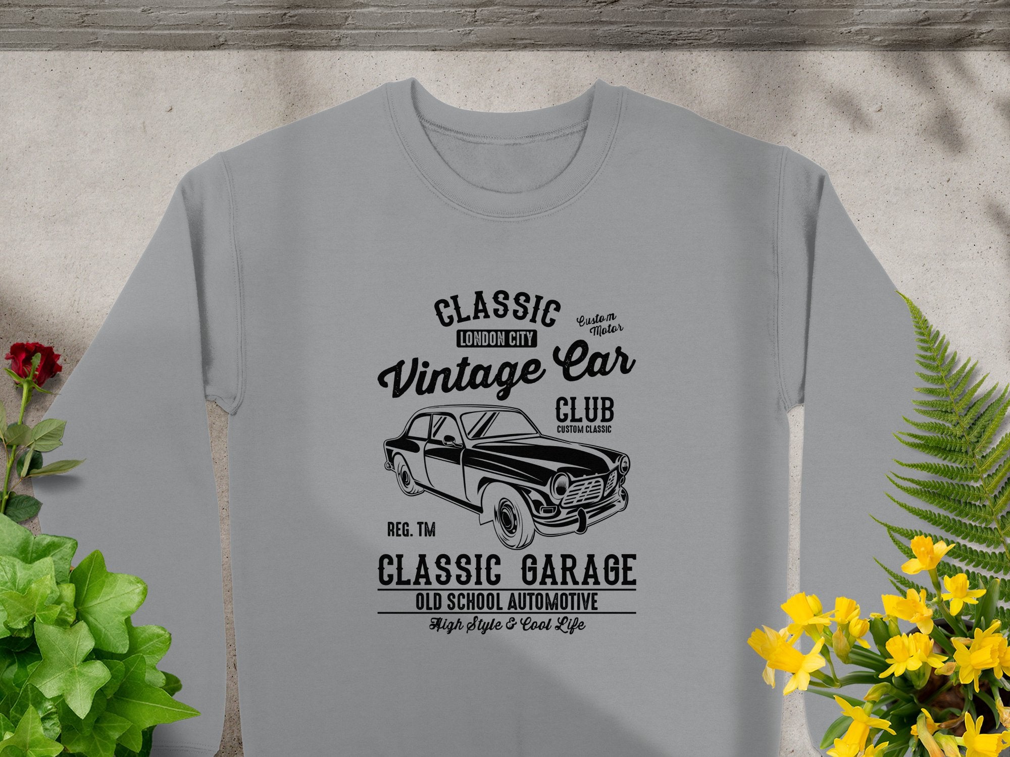 A Garment Graphics gray T-shirt featuring a retro car graphic and text Classic London City Vintage Car Club Custom Garage with High Style & Cool Life is displayed on a flat surface, accented by yellow flowers and green leaves to highlight its automotive flair.
