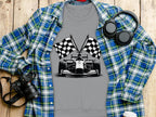 Garment Graphics gray t-shirt with a black and white race car and checkered flag design on a wooden surface, surrounded by a blue and green plaid shirt, camera, headphones, and coffee cup.