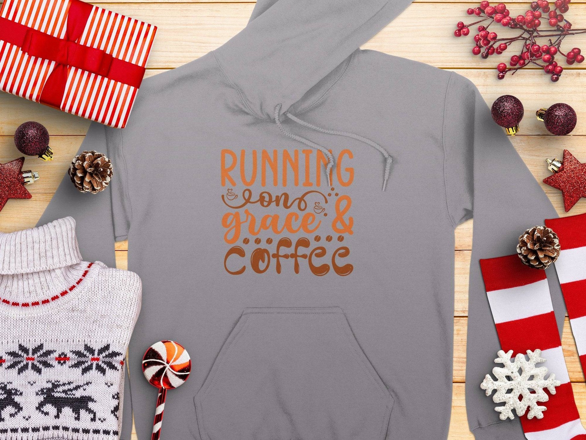 The Garment Graphics Running on Grace and Coffee Hoodie, in a medium-heavy classic fit, displays RUNNING ON GRACE & COFFEE in orange and brown. Its beautifully presented with wrapped gifts, pine cones, and reindeer sweaters on a rustic wooden background.