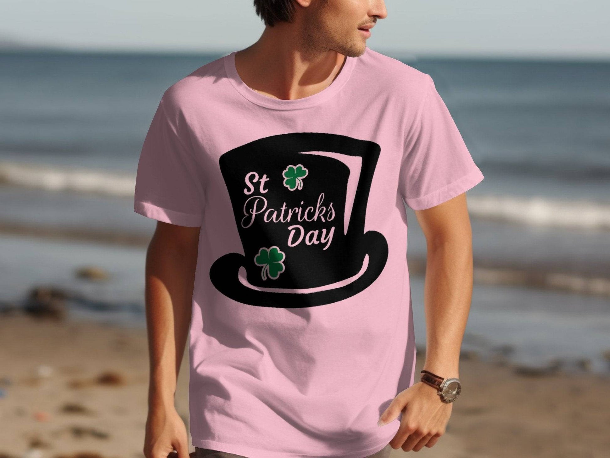 A person walks on the beach wearing a Garment Graphics pink T-shirt featuring a large black top hat with the text St Patricks Day, adorned with two green shamrocks. The serene ocean backdrop enhances this festive attire.