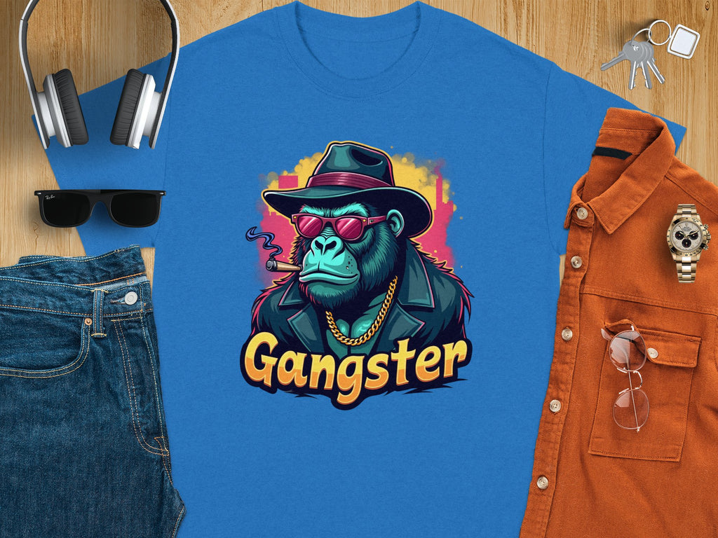 A flat lay features the Garment Graphics Gangster Gorilla T-Shirt, displaying urban style with a gorilla graphic in a hat, sunglasses, and cigar. Surrounding items include headphones, sunglasses, jeans, a watch, keys, and an orange classic fit shirt.