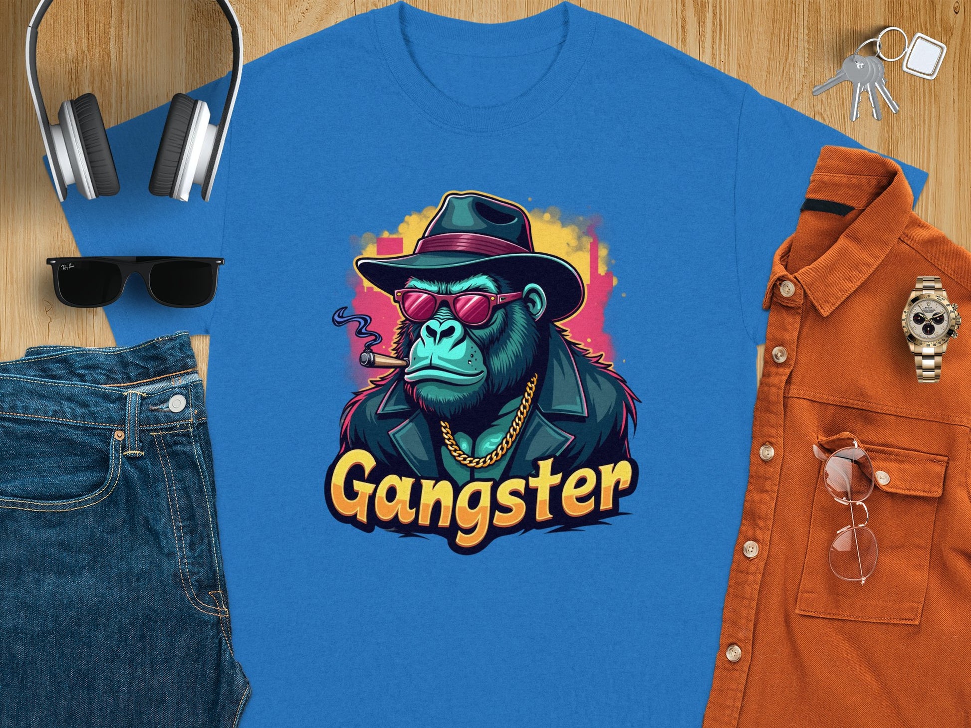 A flat lay features the Garment Graphics Gangster Gorilla T-Shirt, displaying urban style with a gorilla graphic in a hat, sunglasses, and cigar. Surrounding items include headphones, sunglasses, jeans, a watch, keys, and an orange classic fit shirt.