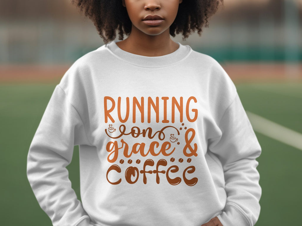 A person wearing a Garment Graphics sweatshirt with Running on Grace & Coffee in brown script stands on a sports field. The unisex sizing ensures comfort for all, with an artfully blurred background.