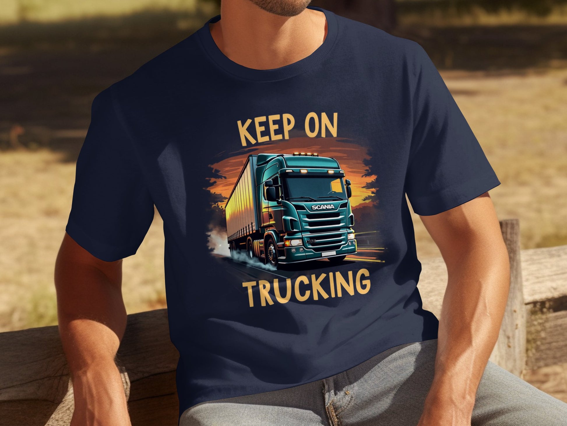 A person in a Garment Graphics navy blue graphic t-shirt showcasing a semi-truck and Keep On Trucking slogan sits on a wooden bench outdoors, surrounded by lush foliage. Perfect for truck enthusiasts.