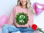Wearing a pink Garment Graphics sweatshirt adorned with a lucky four-leaf clover and surrounded by green clovers, an individual sits cross-legged, holding a red rose beside pink heart-shaped balloons. Their long, wavy hair flows gracefully, embodying the Irish spirit.