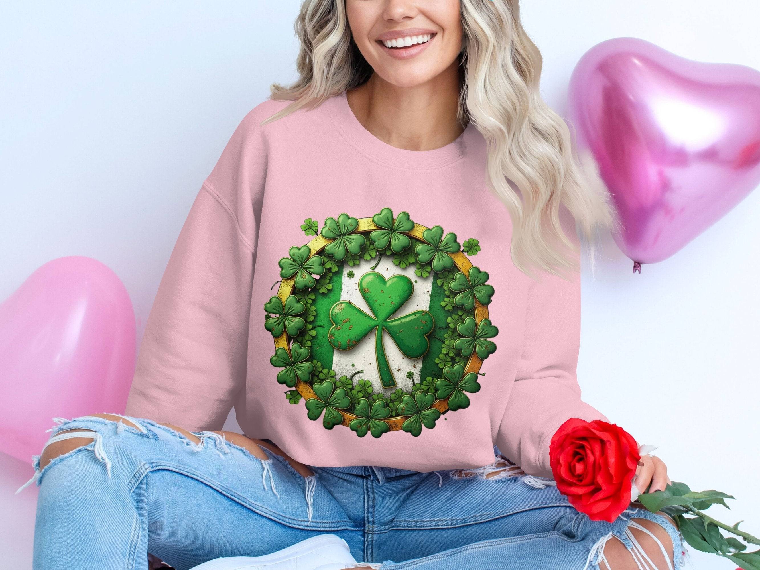 Wearing a pink Garment Graphics sweatshirt adorned with a lucky four-leaf clover and surrounded by green clovers, an individual sits cross-legged, holding a red rose beside pink heart-shaped balloons. Their long, wavy hair flows gracefully, embodying the Irish spirit.