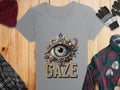 A womens gray Garment Graphics T-shirt with an eye and jewels design and GAZE text lies on wood. Nearby are a red hat, plaid scarf, gloves, plaid shirt, and glasses.