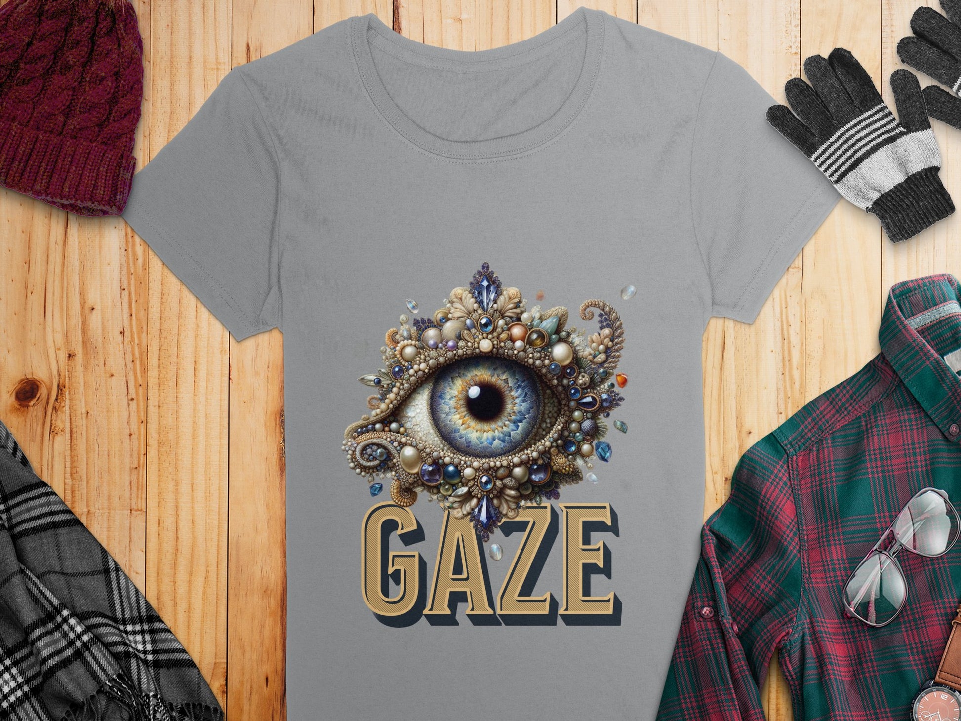 A womens gray Garment Graphics T-shirt with an eye and jewels design and GAZE text lies on wood. Nearby are a red hat, plaid scarf, gloves, plaid shirt, and glasses.