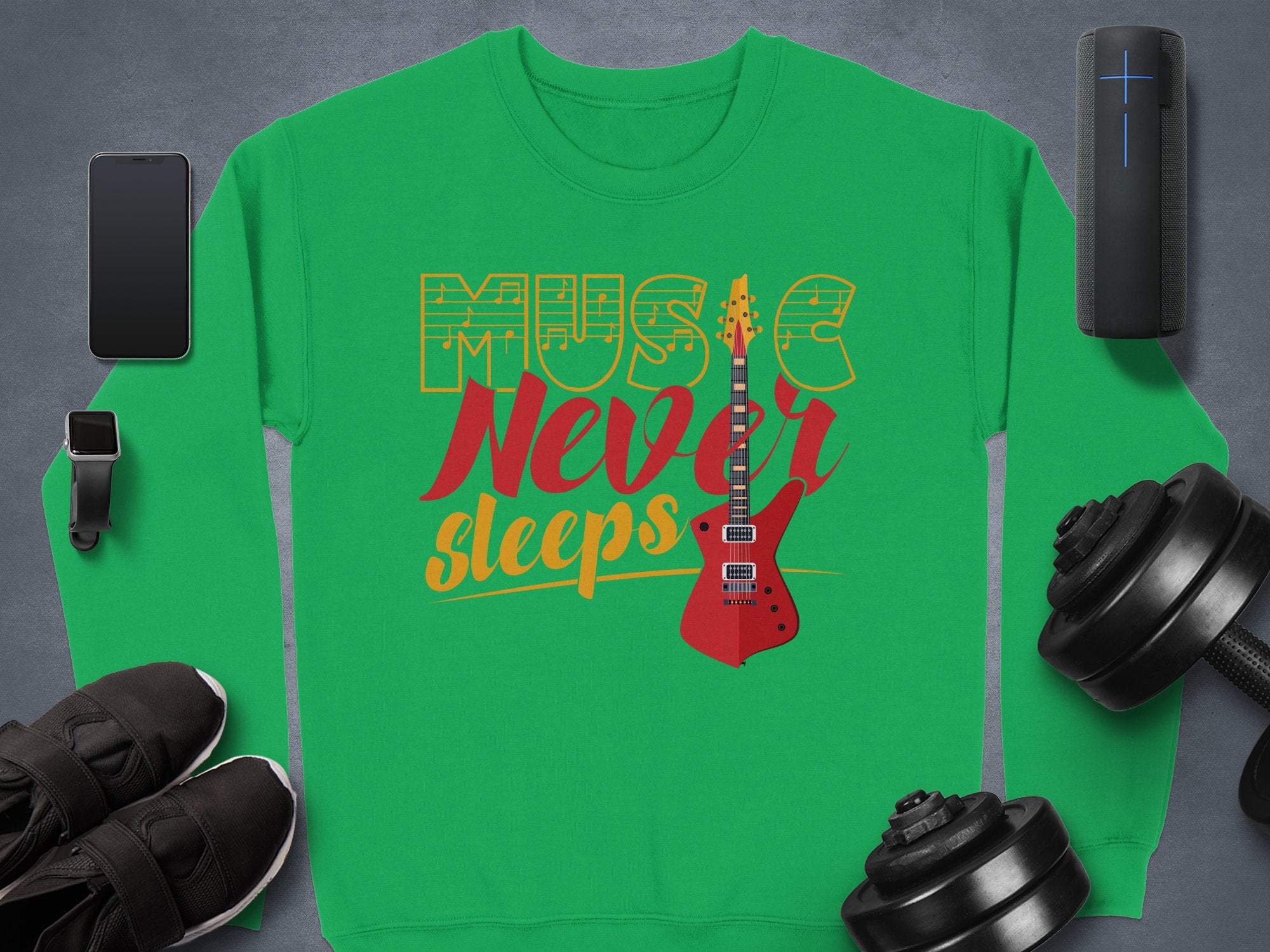 On a gray surface lies a trendy Garment Graphics sweatshirt, featuring Music Never Sleeps and a red electric guitar. Its surrounded by a wireless speaker, dumbbells, black sneakers, a smartwatch, and a smartphone—ideal for music lovers.