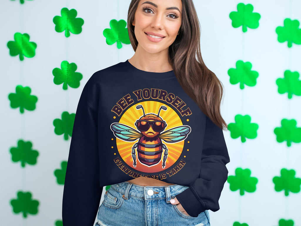 A woman smiles in a dark Bee Yourself Summer Edition sweatshirt from Garment Graphics, standing against a wall with green shamrocks. The unisex sizing means everyone can enjoy this cozy reminder to always be true to oneself.