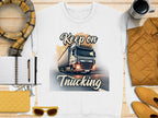 A unisex white graphic sweatshirt from Garment Graphics, showcasing a truck illustration and Keep on Trucking text, is displayed on a wooden surface with a scarf, notebook, pen, yellow accessories, coffee cup, and bright yellow jacket.