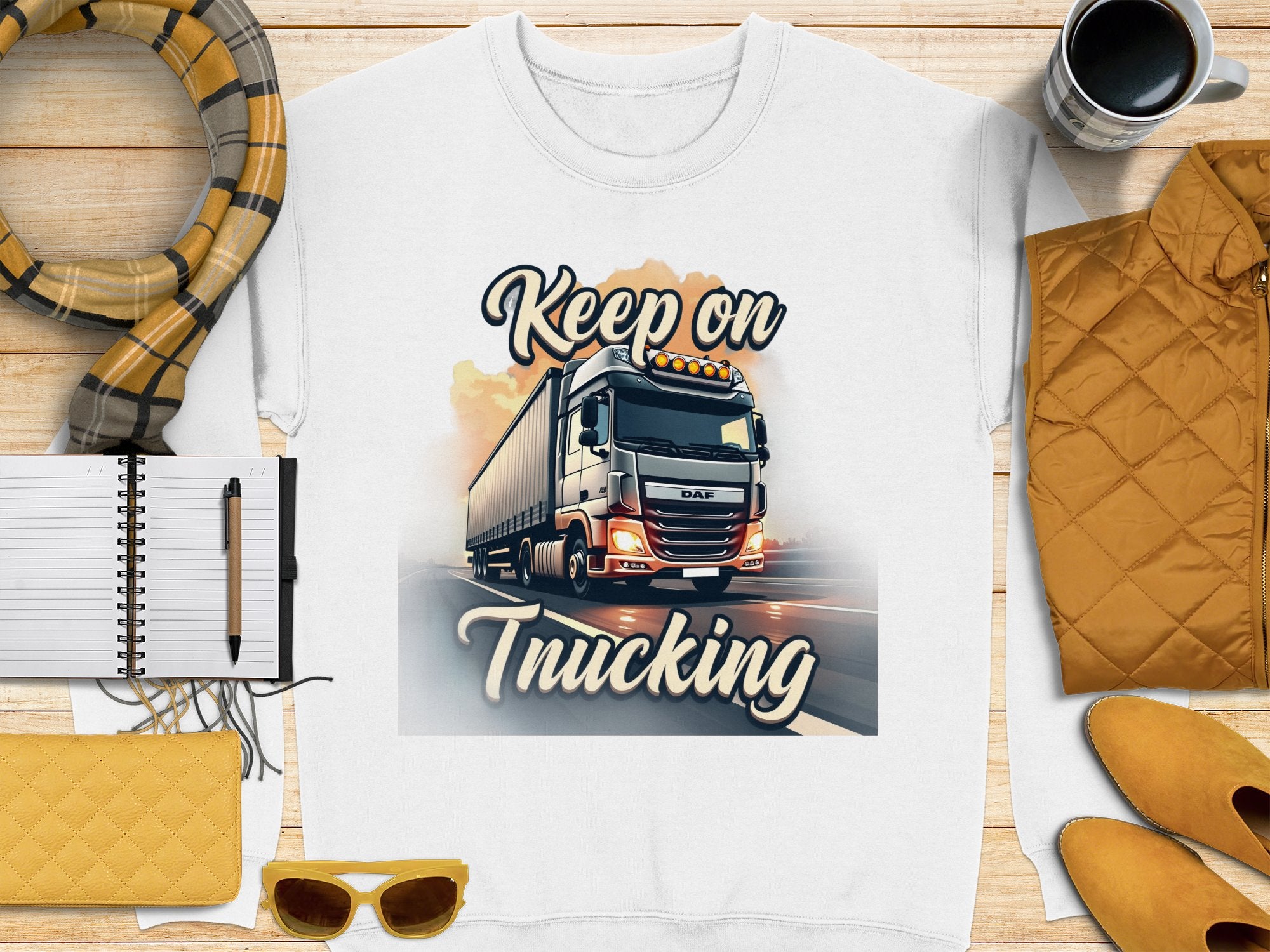 A unisex white graphic sweatshirt from Garment Graphics, showcasing a truck illustration and Keep on Trucking text, is displayed on a wooden surface with a scarf, notebook, pen, yellow accessories, coffee cup, and bright yellow jacket.