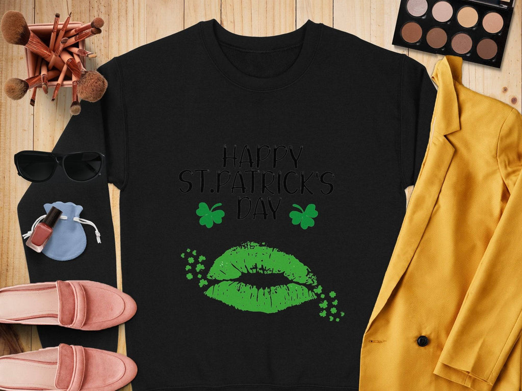 The Garment Graphics black sweatshirt, featuring Happy St. Patricks Day text with green lips and shamrocks, brings a festive vibe. Its styled with makeup brushes, a green eyeshadow palette, sunglasses, a pouch, a mustard blazer, pink shoes, and lipstick on a wooden surface.