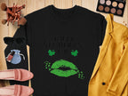 The Garment Graphics black sweatshirt, featuring Happy St. Patricks Day text with green lips and shamrocks, brings a festive vibe. Its styled with makeup brushes, a green eyeshadow palette, sunglasses, a pouch, a mustard blazer, pink shoes, and lipstick on a wooden surface.