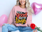 A person with long blonde hair sits on the floor wearing Garment Graphics unisex pink graphic sweatshirt reading Its a good day to laugh. They hold a red rose, surrounded by pink heart balloons. Their ripped jeans pair well with the cozy medium-heavy fabric of the sweatshirt.