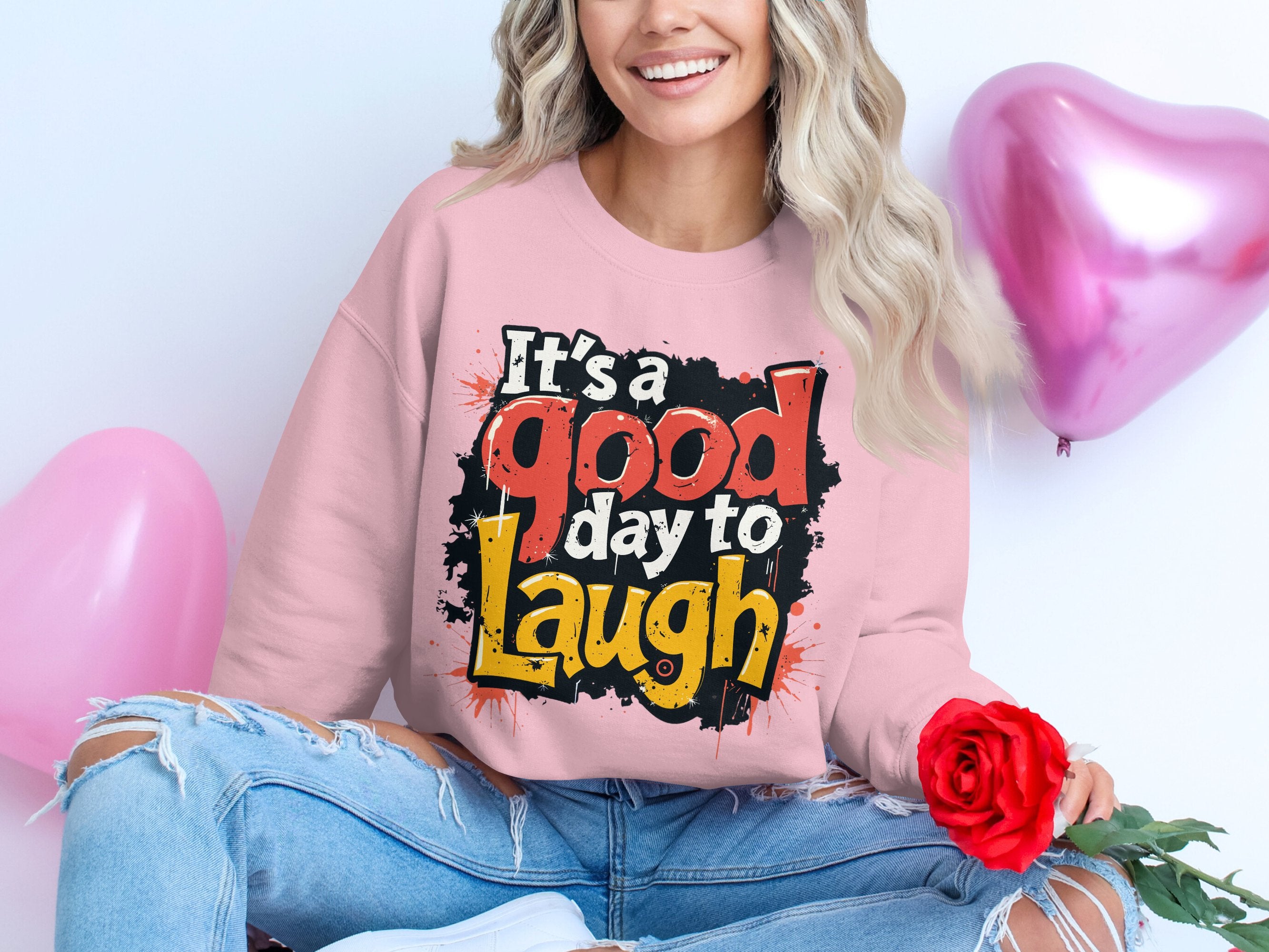 A person with long blonde hair sits on the floor wearing Garment Graphics unisex pink graphic sweatshirt reading Its a good day to laugh. They hold a red rose, surrounded by pink heart balloons. Their ripped jeans pair well with the cozy medium-heavy fabric of the sweatshirt.
