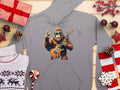 The Garment Graphics electric guitar hoodie features a Bigfoot-like creature rocking out with sunglasses and a peace sign, perfect for music lovers. The design includes wrapped gifts, pine cones, red stars, knit sweater elements, and candy decorations on wood—a must-have for cool vibes!.