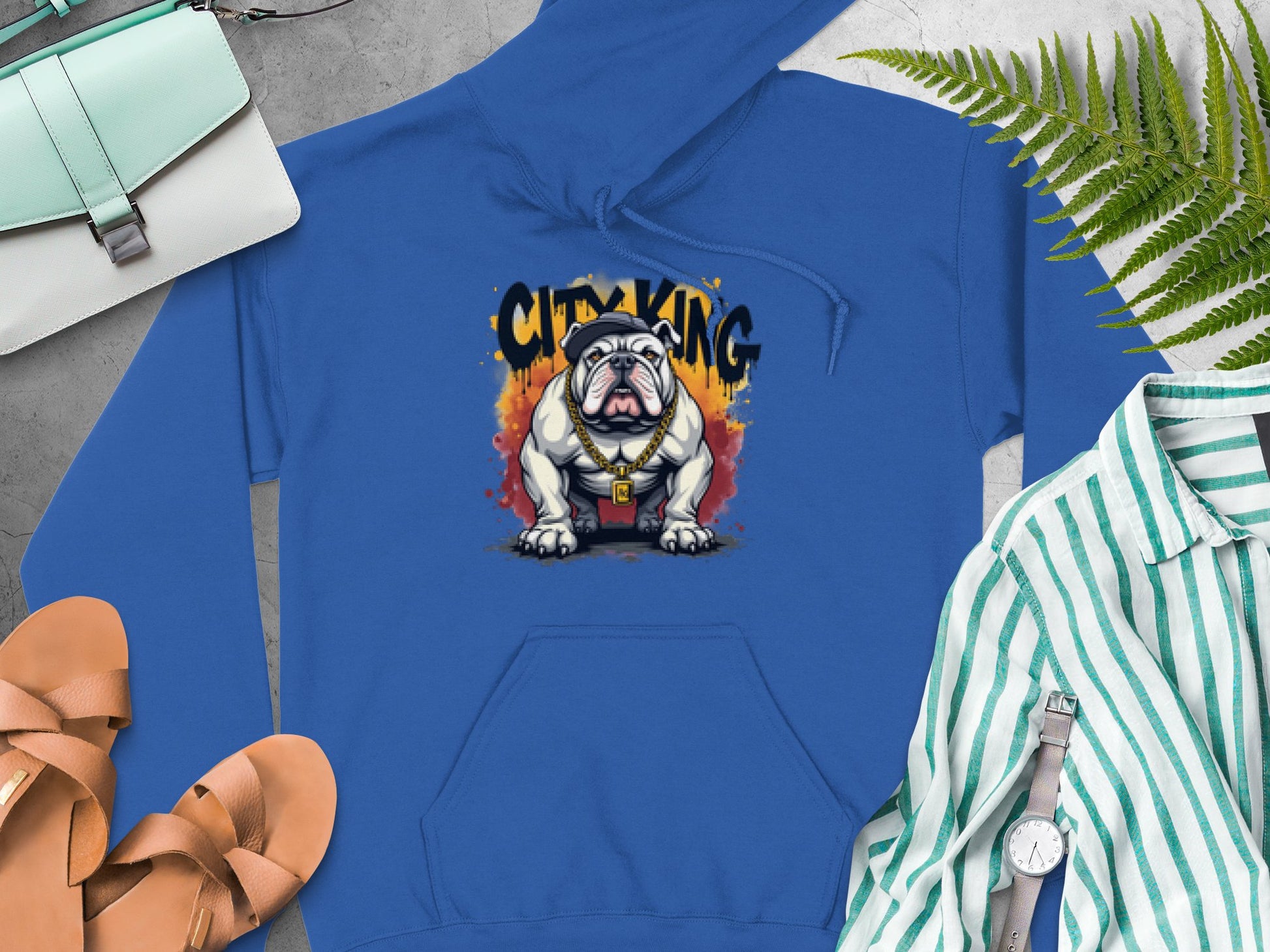 Garment Graphics presents a blue hoodie featuring a bulldog and City King text for urban edge. Displayed on concrete, it is styled with sandals, a striped shirt, handbag options, a watch, and fern leaves.