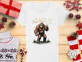 A Garment Graphics white T-shirt features a fun brown Bigfoot peace sign design, labeled Admit It Bigfoot, with the phrase It Would Be Boring Without Me, surrounded by gifts, pine cones, and holiday decor on a wooden surface.