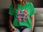 A woman sits on a sofa in a vibrant Garment Graphics 100% cotton green t-shirt featuring colorful balloons and the text Happy Birthday. Casually dressed with her hair down, she rests her hands on her lap, embracing the joyful birthday ambiance.