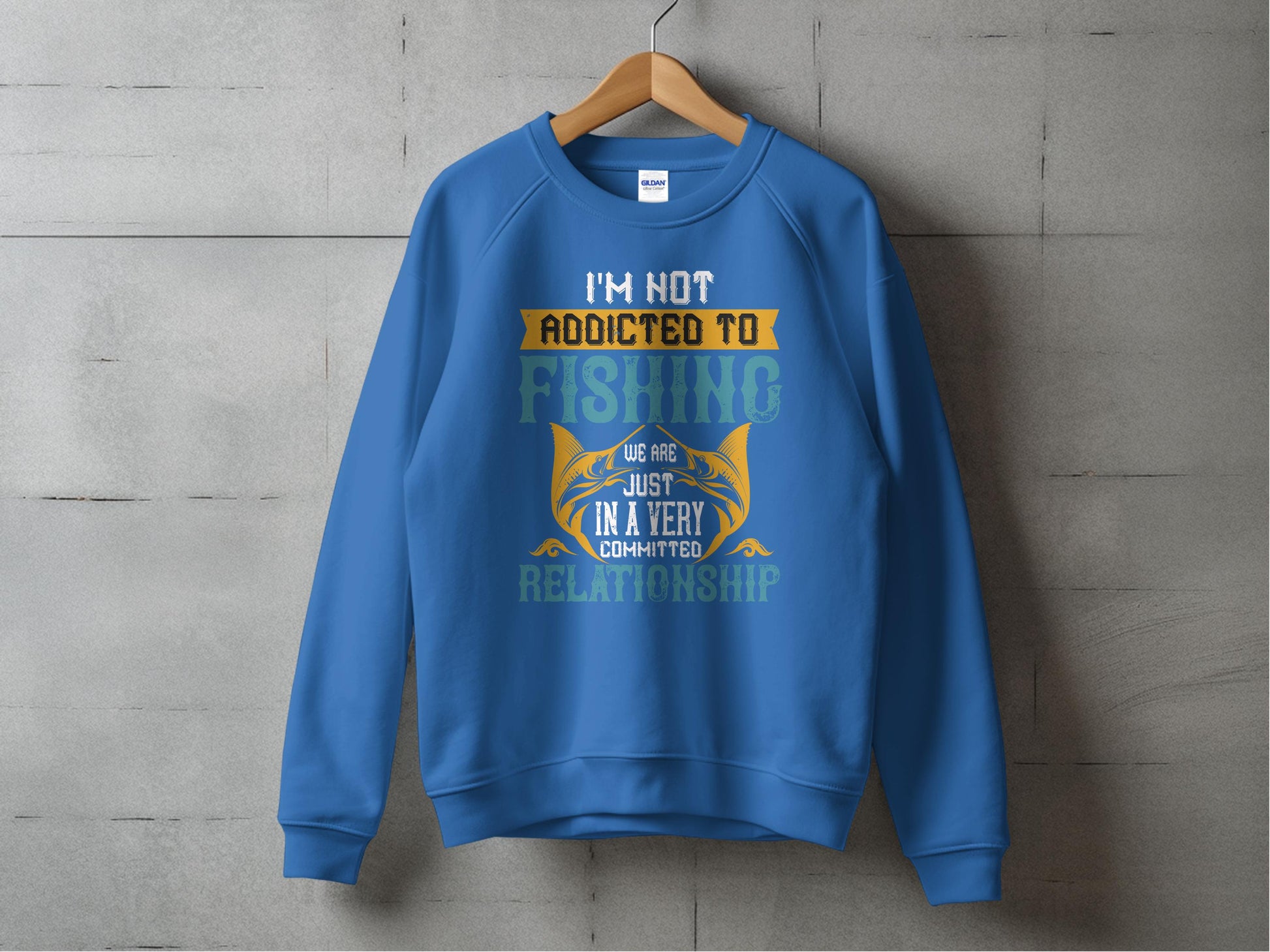 A blue unisex sweatshirt by Garment Graphics hangs on a concrete wall, showcasing the text: Im not addicted to fishing. We are just in a very committed relationship, complete with fishing-themed graphics.