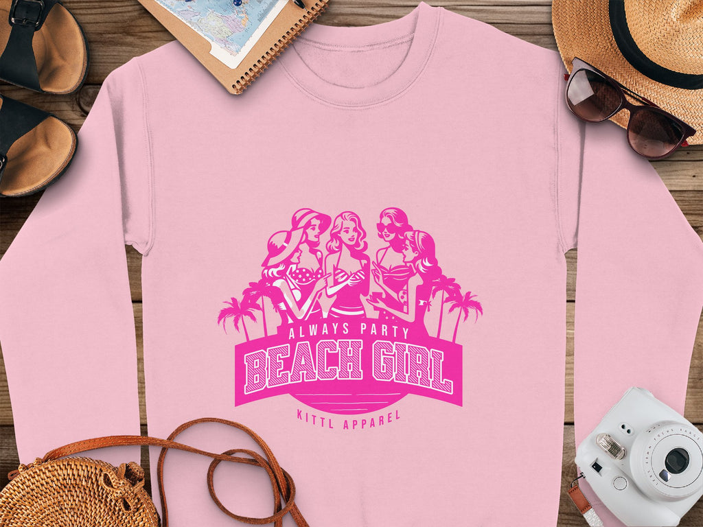 A pink sweatshirt from Garment Graphics is on a wooden surface, showcasing a graphic of three women with palm trees and the text Always Party Beach Girl, surrounded by summer essentials: a straw hat, sunglasses, camera, shoes, and a notebook.