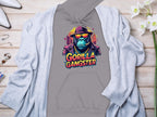 Garment Graphics presents a gray hoodie with vibrant gorilla artwork, featuring a cartoon gorilla in a fedora, sunglasses, and a gold chain with Gorilla Gangster in colorful letters. This streetwear piece is displayed on wood next to a gray cardigan, white shoes, and decorative items.